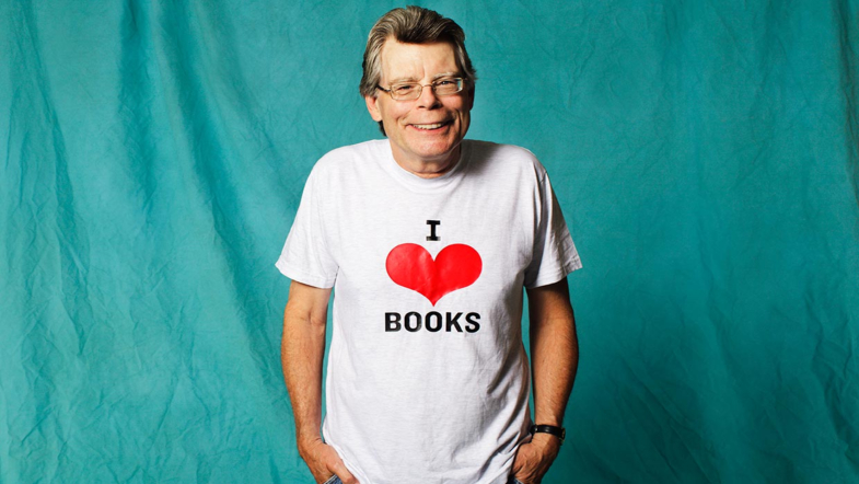 King smiling against blue backdrop with an I heart books shirt on. Hands in pockets.