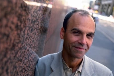 Scott Turow smiling looking at camera, leaning against pink granite wall, wearing grey suit jacket