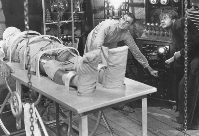 Victor Frankenstein and Fritz standing over the Monster on the slab, preparing to give it life