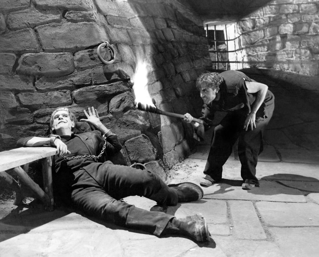 Fritz, played by Dwight Fyre, threatens the Monster, played by Boris Karloff with a burning torch