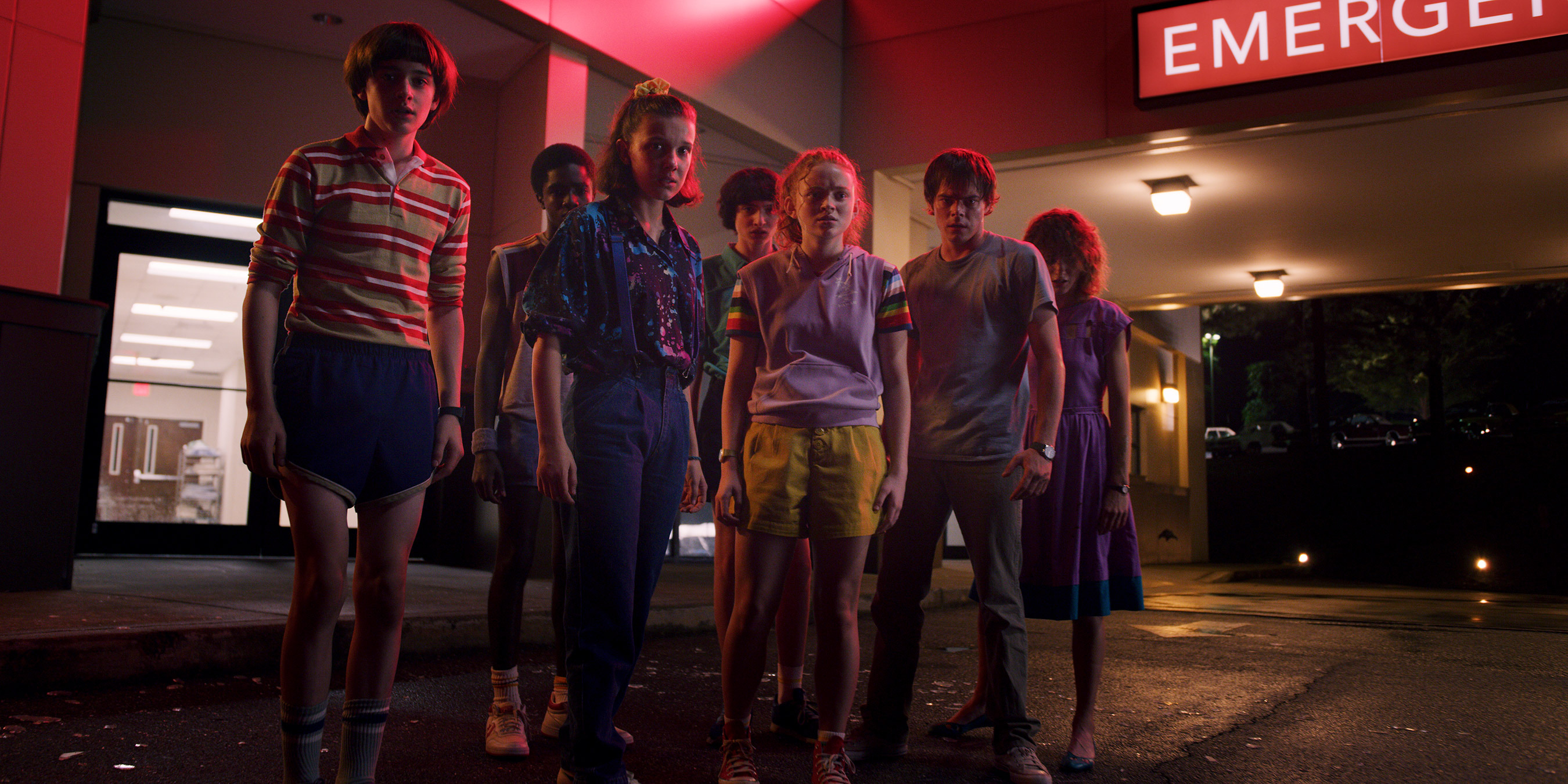 The Stranger Things cast stands assembled in front of a mall, backlit by neon red signs