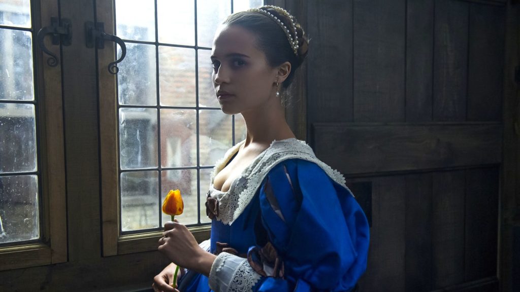 A woman in a period dress stands before a window with a rose