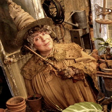 Pomona Sprout stands in a greenhouse clad in Herbology attire with a witch's hat on