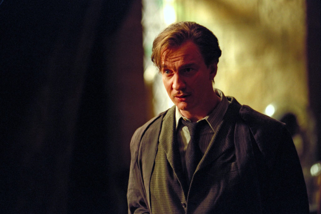 The dapper Remus Lupin stands in a well lit room in a suit and tie 