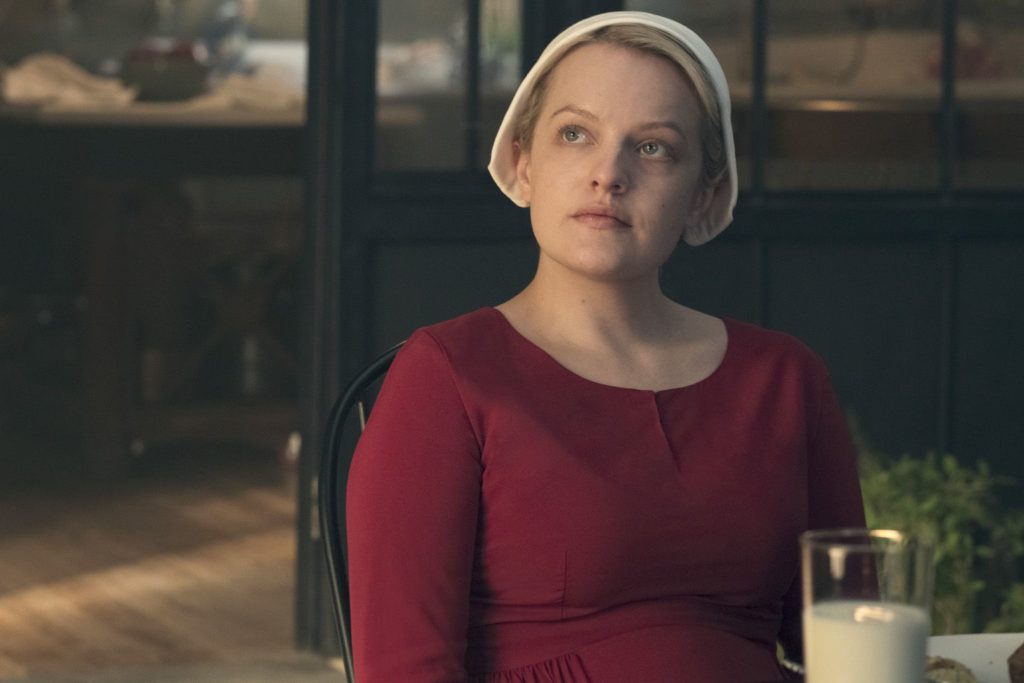 Offred from the Handmaid's Tale sits in a red dress with a table and bottle of milk before her