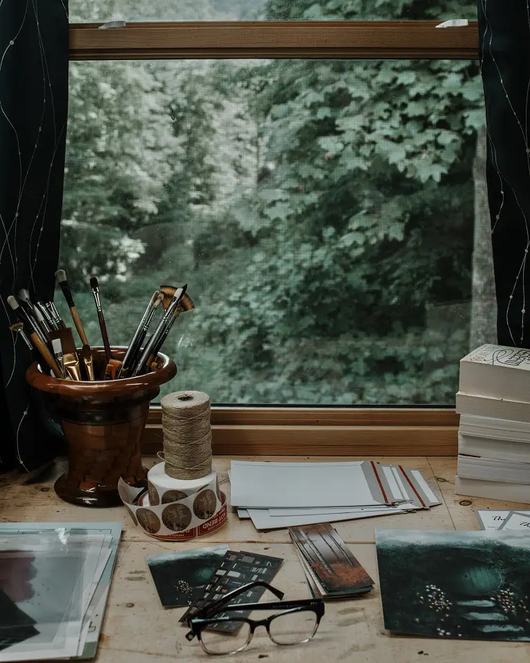 Image of a desk with writing utensils and paper on in with a window in the background that shows a forest