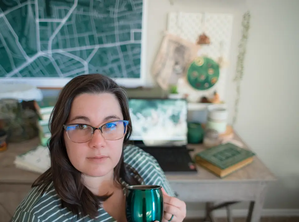 Selfie of a woman holding a mug in front of a desk with a laptop and flower on it