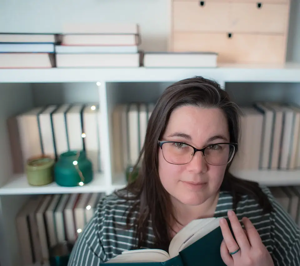 Woman standing in front of a bookcase looking at the camera holding an open book