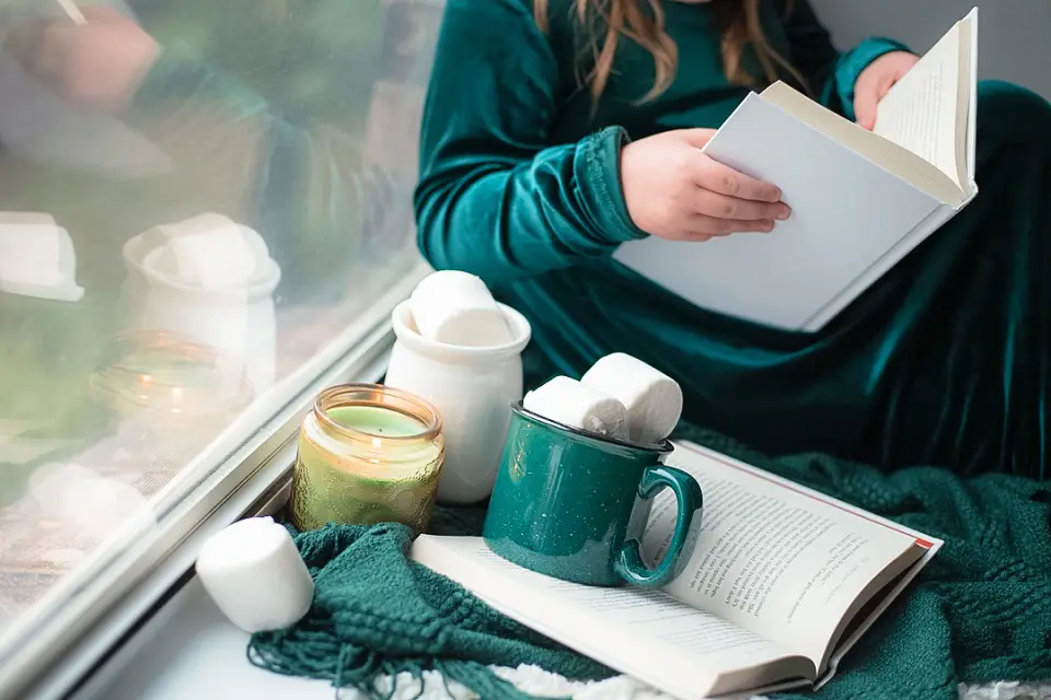 Image centers on sevearl mugs of coffee with a woman sitting next to them reading a book