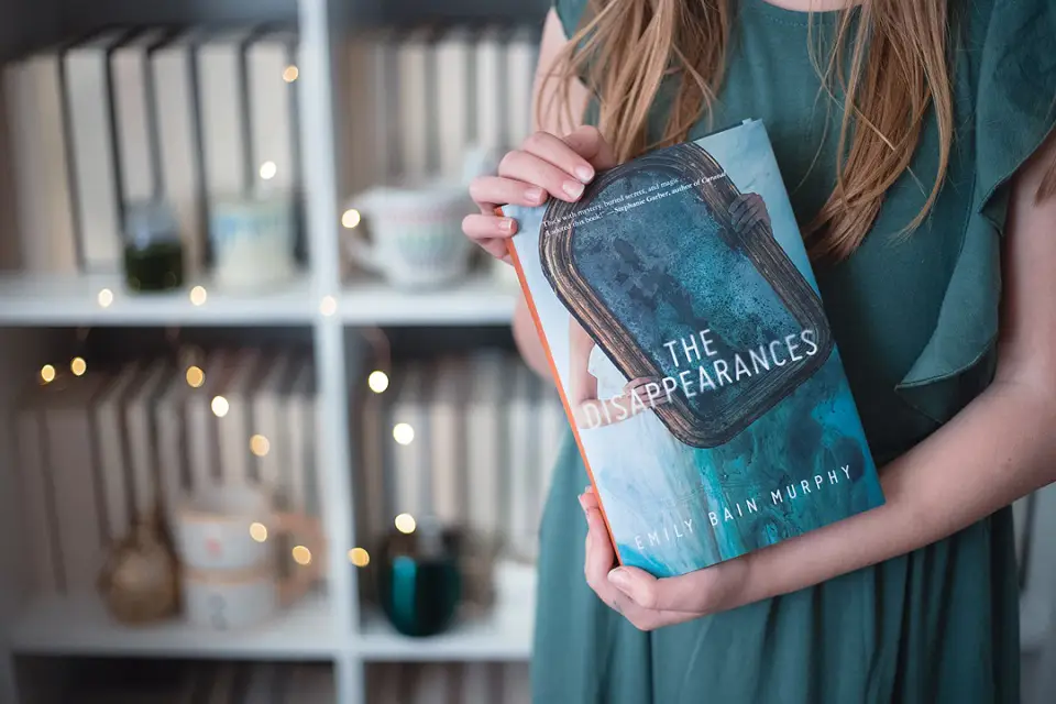 Half shot of a woman holding a book showing the camera the cover in front of a bookshelf