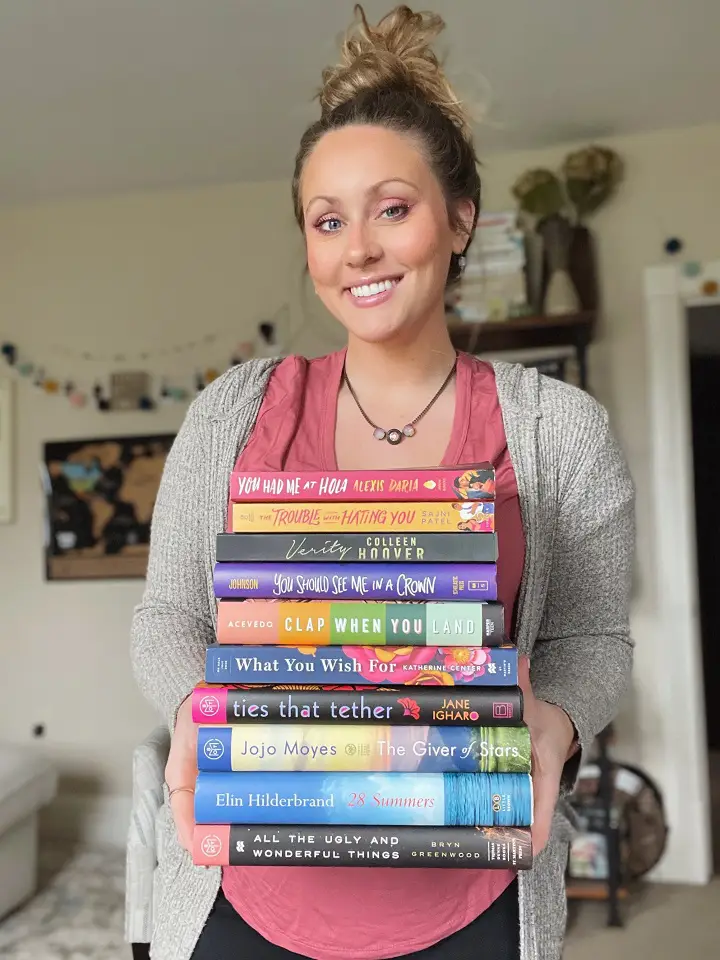 A woman in a oink shirt and gray sweater holding a stack of books smiling for the camera