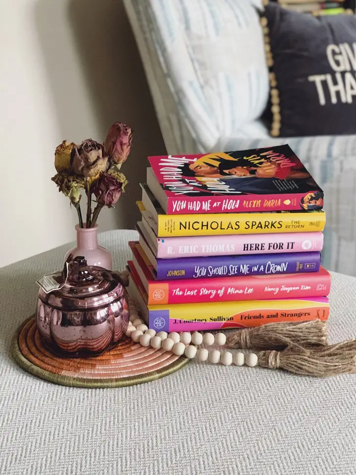 A stack of books on a table next to a vase with a shriveled flower 
